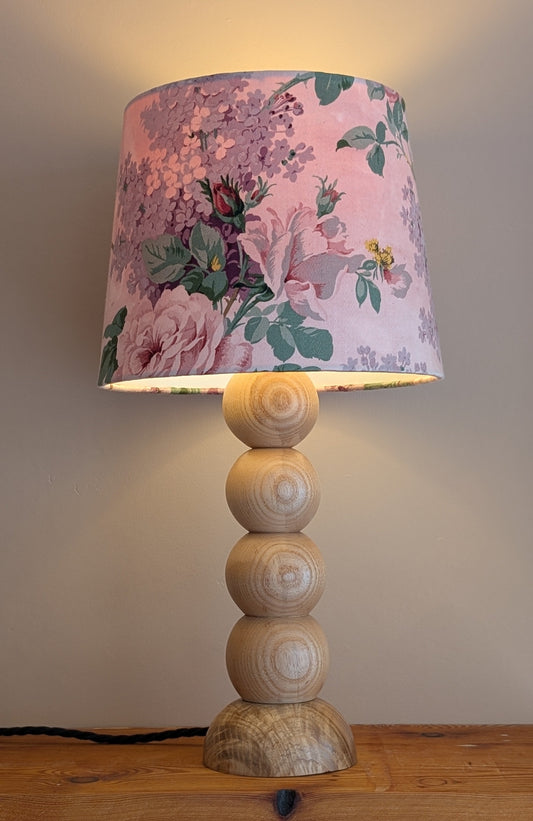 A table lamp with a pink floral patterned drum shade and a stacked wooden base, shown lit on a wooden shelf against a plain wall.