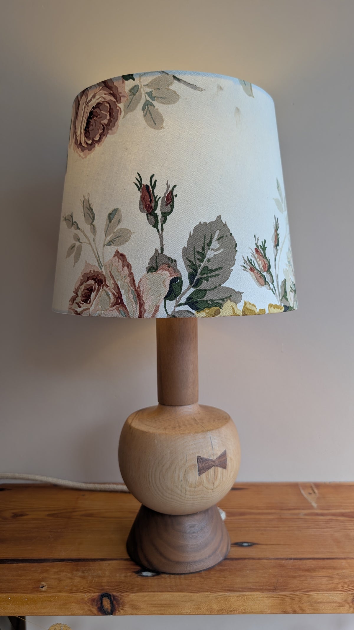 A table lamp with a light natural wood ball-shaped base and a tapered drum shade featuring a vintage floral pattern in muted pink, green, and brown tones, set on a wooden surface against a plain wall.