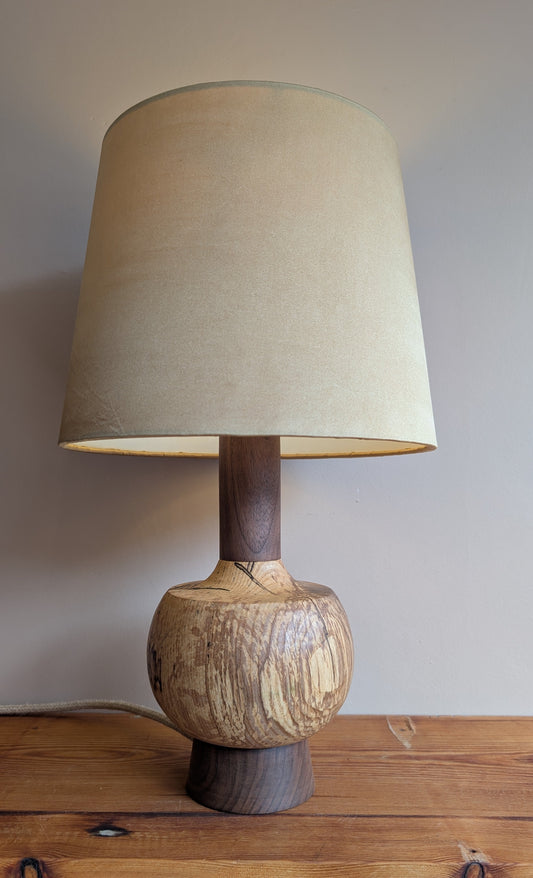 A wooden table lamp with a tapered beige fabric shade, a dark wood neck, and a round carved wooden base, shown on a wooden shelf against a plain wall.